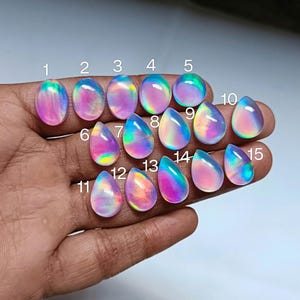 Op de afbeelding: Vijftien regenboogkleurige, traanvormige cabochons in verschillende tinten roze, blauw en paars. De cabochons zijn in rijen op een hand gerangschikt.