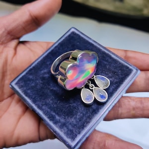 May include: A silver ring featuring a cloud-shaped iridescent stone and three dangling teardrop-shaped stones. The ring is presented in a blue velvet box.