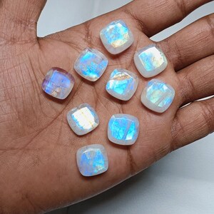 Peut inclure: Une collection de dix cabochons de pierre de lune blanche iris&eacute;e, chacun avec un motif unique de reflets bleus et verts. Les pierres sont taill&eacute;es en forme carr&eacute;e avec des bords arrondis et sont dispos&eacute;es dans une main.