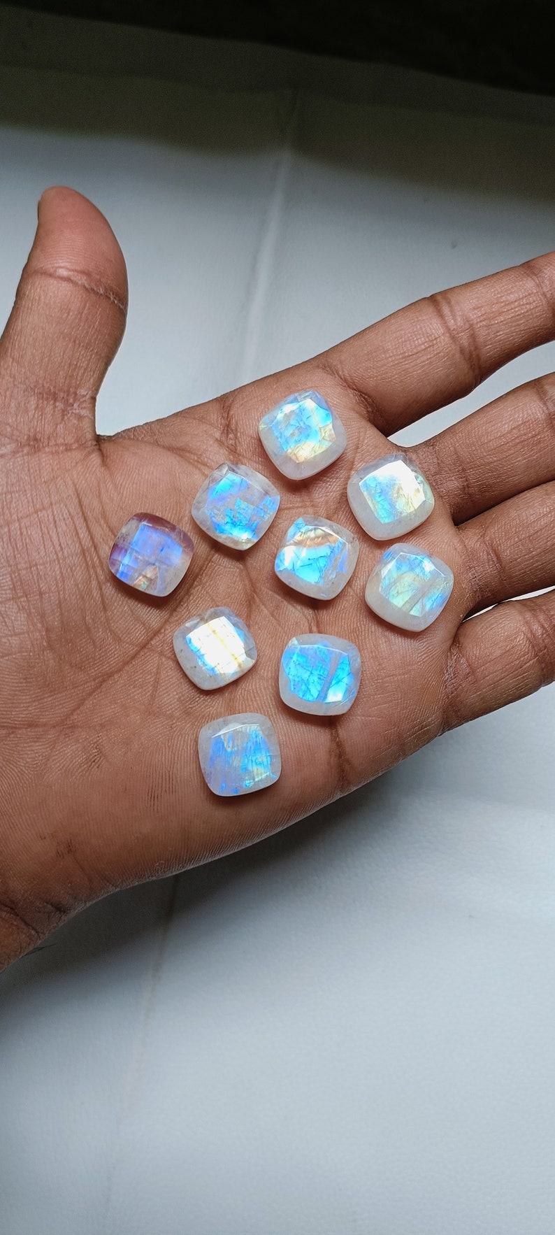 Peut inclure: Une collection de dix cabochons de pierre de lune blanche iris&eacute;e, chacun avec un motif unique de reflets bleus et verts. Les pierres sont taill&eacute;es en forme carr&eacute;e avec des bords arrondis et sont dispos&eacute;es dans une main.