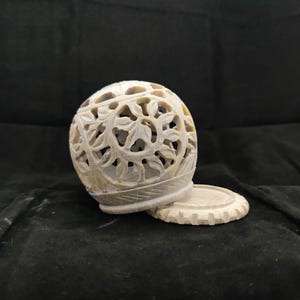 Peut inclure: Une sphère en pierre sculptée avec des motifs complexes de feuilles et de vignes, posée sur une base circulaire assortie. La sphère est blanc cassé avec de subtiles variations de couleur. Un objet décoratif.