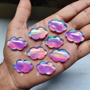 May include: Nine iridescent cloud-shaped gemstones in shades of purple, pink, and blue. Each stone has a unique color pattern and is displayed on a hand. The gemstones are likely for jewelry making or crafting.
