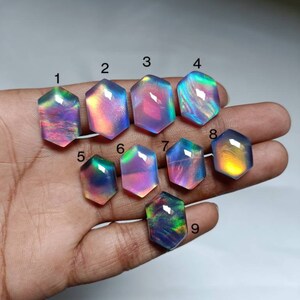 Puede incluir: Nueve cabujones hexagonales iridiscentes en varios tonos de morado, azul y verde. Los cabujones están dispuestos en una fila en una mano.