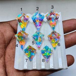 Puede incluir: Ocho piedras de vidrio transparente facetadas e iridiscentes con colores arcoíris. Las piedras están dispuestas en filas sobre una superficie blanca.