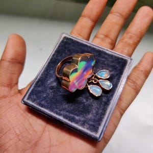 May include: A gold-toned ring with a large, iridescent, flower-shaped stone. Three smaller, teardrop-shaped stones dangle below the flower. The ring is displayed on a dark blue velvet surface inside a clear plastic case.
