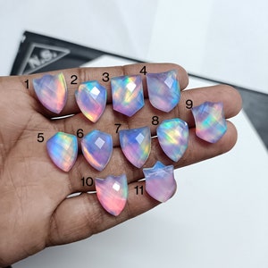 May include: Eleven shield-shaped, faceted opal-like gemstones with iridescent, translucent pink and blue hues. Each stone is numbered from 1 to 11, displaying a polished surface that reflects light with a multi-coloured, glowing effect.