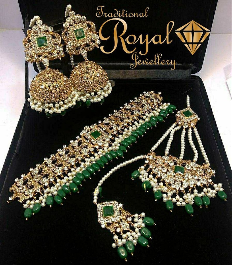 Op de afbeelding: Een traditionele Indiase bruidssieradenset met een gouden chokerketting met groene en witte kralen, een bijpassende maang tikka en een paar jhumka-oorringen. De sieraden zijn versierd met glinsterende stenen en parels.