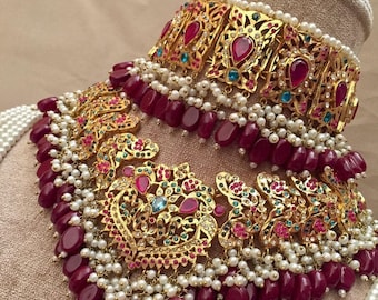 Op de afbeelding: Een gouden kettingset met ingewikkelde bloemmotieven, bezet met rode en groene edelstenen en witte parels. De ketting heeft een gelaagd ontwerp met een choker-stijl bovenstuk en een langer onderstuk.