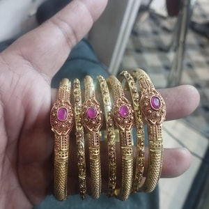 May include: A set of six gold bangles with intricate designs and pink gemstones. The bangles are stacked on top of each other.