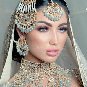 Könnte beinhalten: Eine Frau in einem traditionellen indischen Brautkleid mit einem weißen Schleier und einem silbernen und goldenen Kopfschmuck mit Perlen. Sie trägt auch eine silberne und goldene Halskette mit Perlen und ein passendes Paar Ohrringe.