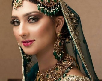 Op de afbeelding: Een vrouw in een groene en gouden bruidsjurk met ingewikkelde borduursels en versieringen. Ze draagt ​​een traditionele Indiase hoofdtooi met parels en groene stenen, en bijpassende oorbellen en een ketting.
