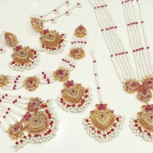 May include: A traditional Indian bridal jewellery set featuring a long pearl and red bead necklace, matching earrings, and a headpiece. The set is adorned with gold accents and pink gemstones.
