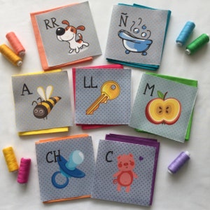 以下が含まれることがあります： 異なる文字と画像が描かれた、カラフルなフェルトの正方形10枚セット。文字はA、CH、C、LL、M、N、RR。画像は、ミツバチ、おしゃぶり、ブタ、鍵、リンゴ、バスタブ、犬です。