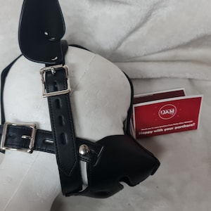 May include: Black leather dog muzzle with a strap and buckle. The muzzle has a nose piece and two ear pieces. The strap is adjustable and has a buckle closure. The muzzle is on a white surface.