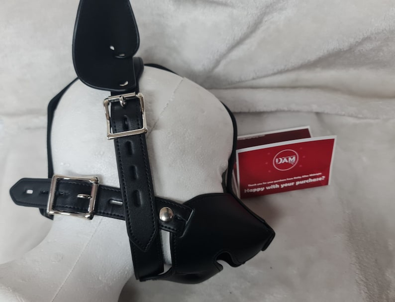 May include: Black leather dog muzzle with a strap and buckle. The muzzle has a nose piece and two ear pieces. The strap is adjustable and has a buckle closure. The muzzle is on a white surface.
