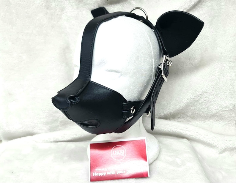 May include: Black leather dog muzzle with a strap and ear pieces. The muzzle has a nose and mouth opening. The strap is adjustable and has a silver buckle. The ear pieces are attached to the strap and are shaped like dog ears.