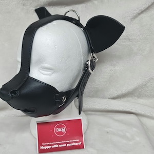 May include: Black leather dog muzzle with ears and a strap. The muzzle has a nose, mouth, and eye holes. The strap is adjustable and has a buckle. The muzzle is on a white mannequin head.