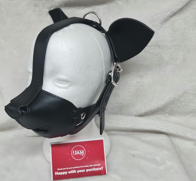 May include: Black leather dog muzzle with ears and a strap. The muzzle has a nose, mouth, and eye holes. The strap is adjustable and has a buckle. The muzzle is on a white mannequin head.