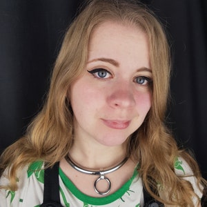 May include: A silver-colored metal collar with a large ring pendant. The collar is worn over a green and white dinosaur-patterned shirt and black overalls. The background is a solid black.