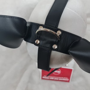 May include: Black leather dog muzzle with two ear flaps and a silver metal ring. The muzzle is on a white background.