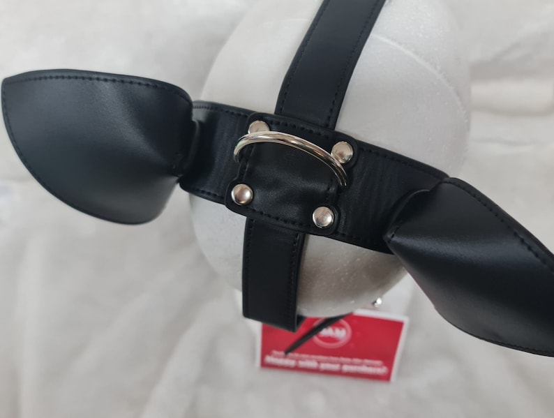 May include: Black leather dog muzzle with two ear flaps and a silver metal ring. The muzzle is on a white background.