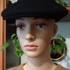 May include: A black wool newsboy cap with a flat top and a curved brim. The cap is worn by a mannequin with a brown sweater.