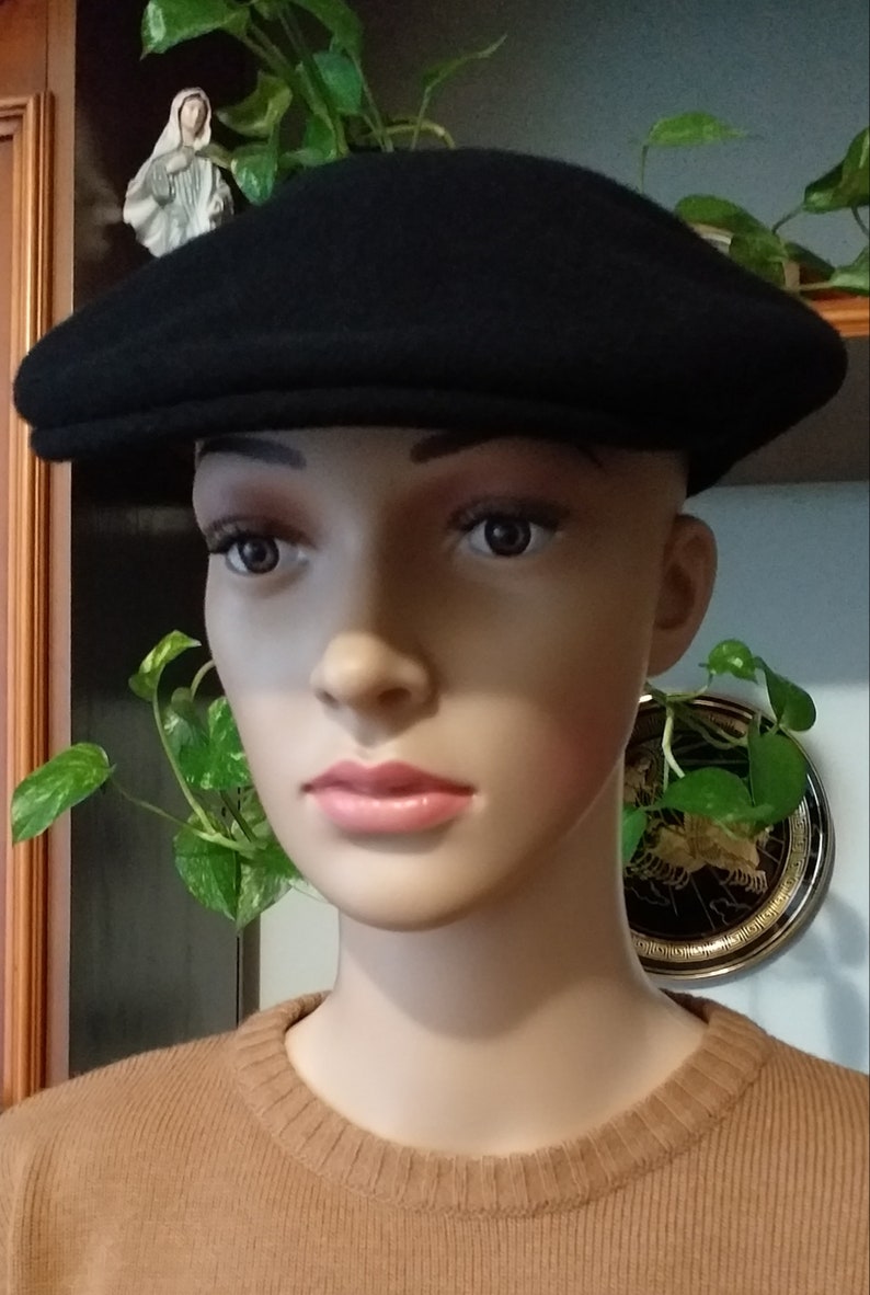 May include: A black wool newsboy cap with a flat top and a curved brim. The cap is worn by a mannequin with a brown sweater.