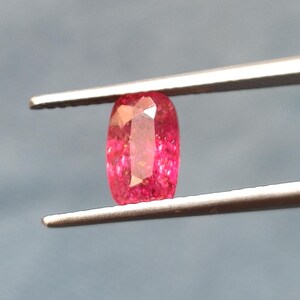 Puede incluir: Una vibrante gema rosa, de corte ovalado, es sostenida por unas pinzas plateadas sobre un fondo gris. Las facetas de la gema capturan la luz, mostrando una gama de tonos rosados. Esta gema probablemente se utiliza en joyería.