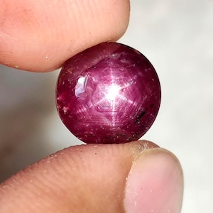 Könnte beinhalten: Ein runder, polierter Rubin-Edelstein, der zwischen zwei Fingern gehalten wird. Der Rubin hat eine tiefe, satte rote Farbe mit einem sternartigen Effekt auf der Oberfläche. Der Edelstein ist glatt und reflektierend.