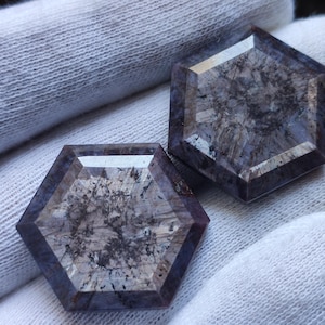 May include: Two hexagonal cut gemstones featuring a translucent center with dark, intricate inclusions and deep purple-toned edges. The stones have a faceted surface that reflects light, showcasing a complex internal pattern of dark mineral deposits.