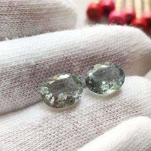 Puede incluir: Dos gemas ovaladas de color verde pálido se muestran sobre una superficie blanca texturizada. Las gemas tienen múltiples facetas, que reflejan la luz. El fondo incluye una disposición borrosa de pequeños objetos rojos y naranjas.