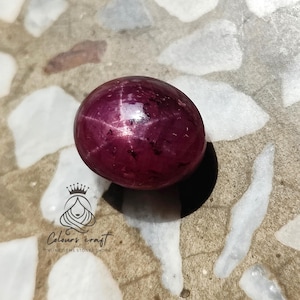 Puede incluir: Una gema pulida, redonda, de color rojo intenso, con un patrón en forma de estrella. La gema está colocada sobre una superficie moteada de color claro. El logotipo de "Colours Craft" es visible en la esquina inferior izquierda.
