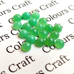 Puede incluir: Un conjunto de gemas redondas y translúcidas de color verde claro. Los cabujones están dispersos sobre una superficie blanca con la inscripción "Colours Craft" en negro. Las gemas son lisas y pulidas.