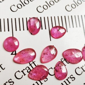 May include: A collection of nine polished, oval and teardrop-shaped, pink gemstones. The gems are arranged on a white surface with a ruler and the word "Craft" visible. The gemstones vary in size and have a translucent appearance.