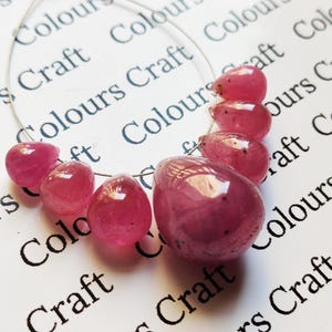 Könnte beinhalten: Strang aus polierten, durchscheinenden, rosa Edelsteinperlen. Die Perlen variieren in Größe und Form und sind auf einem dünnen Draht aufgereiht. Der Hintergrund ist weiß mit den sich wiederholenden Wörtern "Colours Craft".
