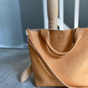 May include: A tan leather tote bag with a long strap and two top handles. The bag has a small logo that reads "Lui-g Bags Handmade Natural".