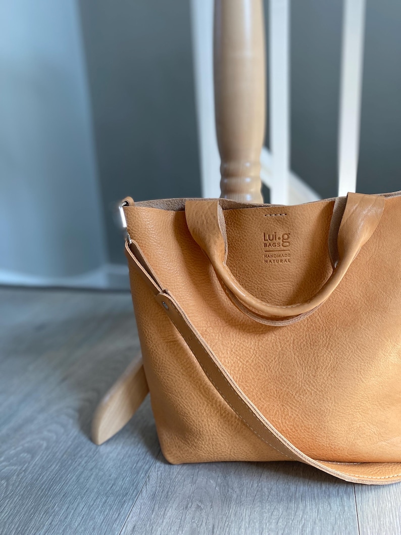May include: A tan leather tote bag with a long strap and two top handles. The bag has a small logo that reads "Lui-g Bags Handmade Natural".