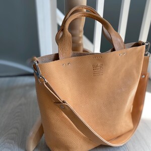 May include: A tan leather tote bag with a long shoulder strap and two top handles. The bag has a small logo that reads "Lui-g Bags Handmade Natural".