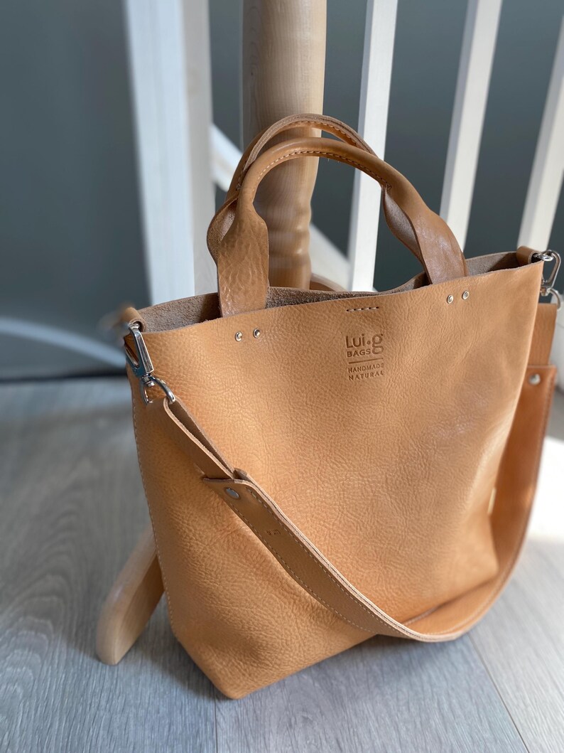 May include: A tan leather tote bag with a long shoulder strap and two top handles. The bag has a small logo that reads "Lui-g Bags Handmade Natural".