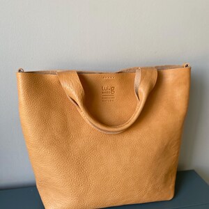 May include: A tan leather tote bag with a top handle and a Luig Bags label that says "Handmade Natural".