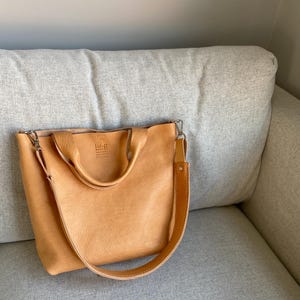 May include: A light brown leather tote bag with a long strap and a smaller top handle. The bag has a logo that says "Lul&ugrave; & Co. Handmade Natural Leather".