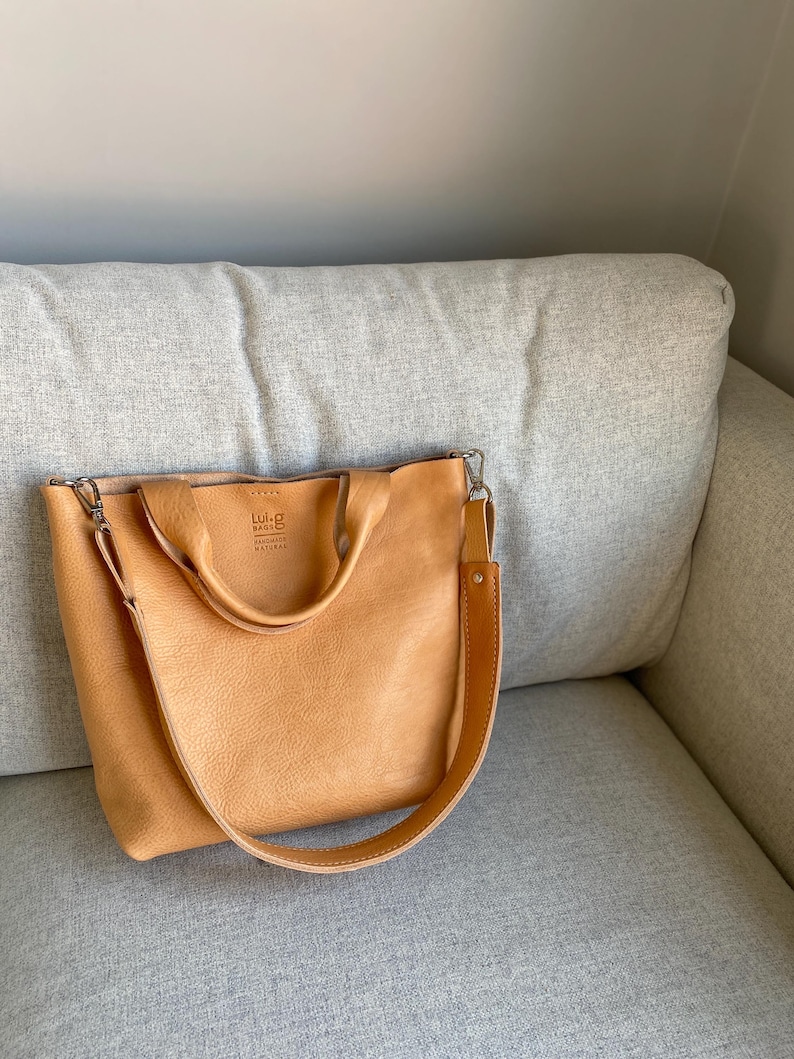 May include: A light brown leather tote bag with a long strap and a smaller top handle. The bag has a logo that says "Lul&ugrave; & Co. Handmade Natural Leather".