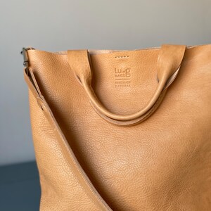 May include: A tan leather tote bag with a long strap and two shorter handles. The bag has a logo that says "Lui-G Bags Handmade Natural".