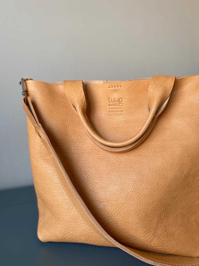 May include: A tan leather tote bag with a long strap and two shorter handles. The bag has a logo that says "Lui-G Bags Handmade Natural".