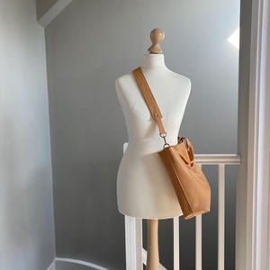 May include: A tan leather tote bag with a long strap hanging from a white mannequin. The bag is on a white railing with a wooden banister.