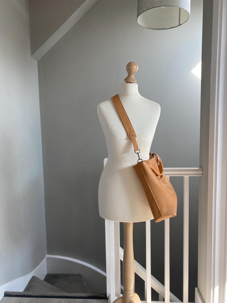 May include: A tan leather tote bag with a long strap hanging from a white mannequin. The bag is on a white railing with a wooden banister.