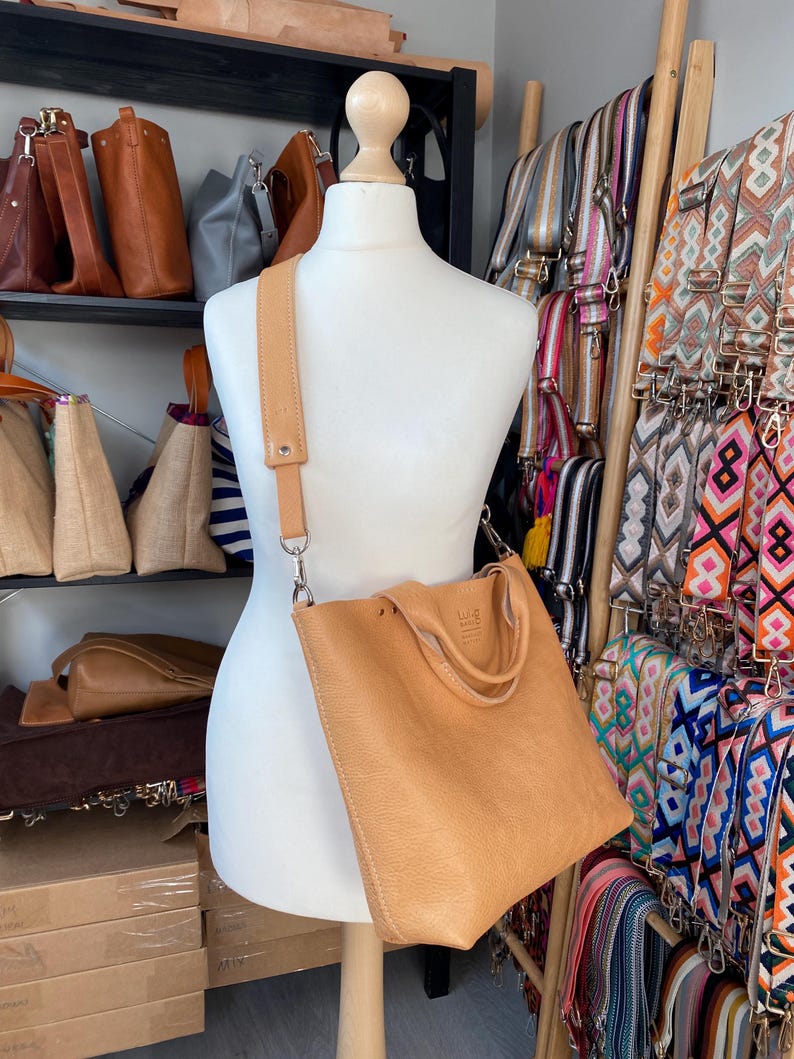 May include: A tan leather tote bag with a long tan leather strap. The bag is on a white mannequin and has the text "LILA MADRID" on the front.