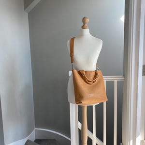 May include: A tan leather tote bag with a long strap, displayed on a wooden mannequin against a white wall.