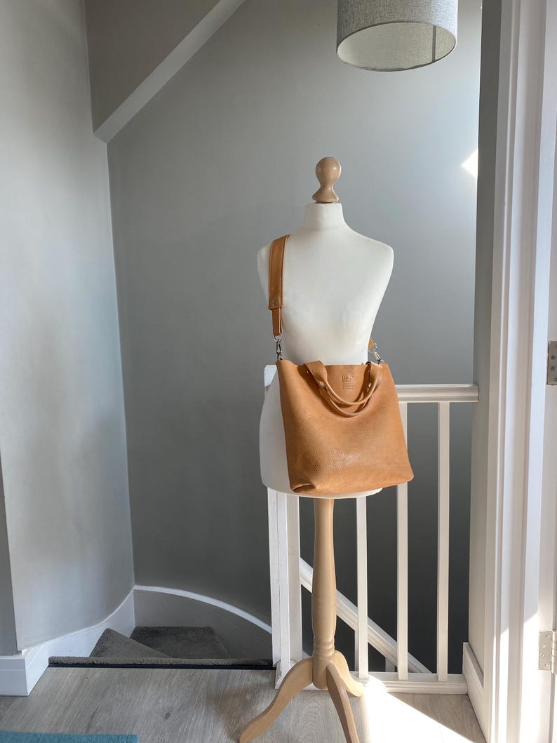 May include: A tan leather tote bag with a long strap, displayed on a wooden mannequin against a white wall.