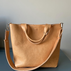 May include: A tan leather tote bag with a long strap and two shorter handles. The bag has a logo that says "Luig Bags Handmade Natural".